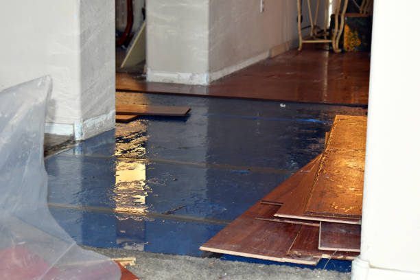 A room with a water-damaged wooden floor and stacked wooden planks on the side. Plastic sheets are visible on the left.